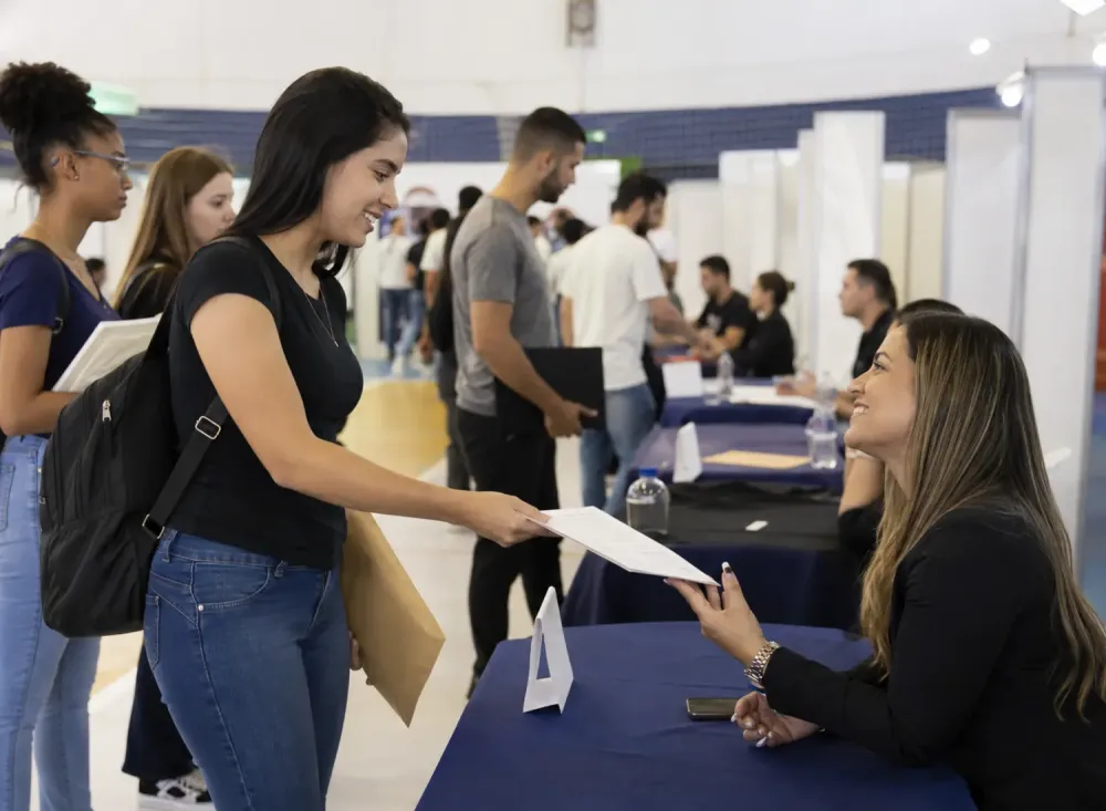  jovem entrega currículo a uma recrutadora durante uma feira de carreiras. Imagem ilustrativa gerada por inteligência artificial/OpenAI.