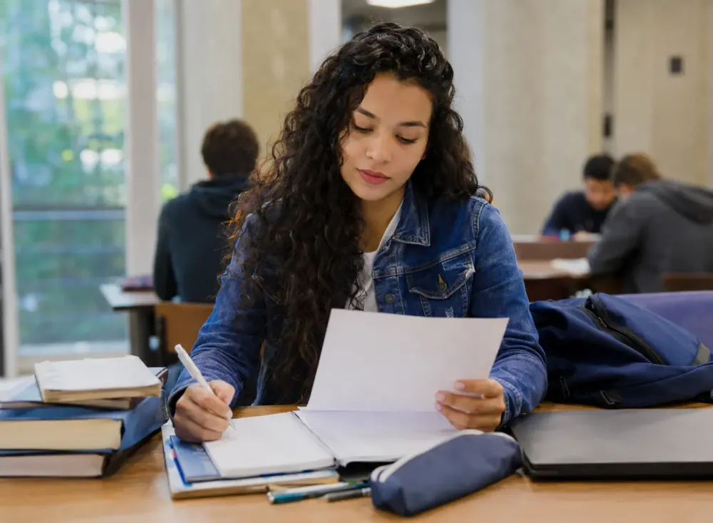  Estudante universitária confere documentos acadêmicos em uma mesa de estudos. Imagem ilustrativa gerada por inteligência artificial/OpenAI.